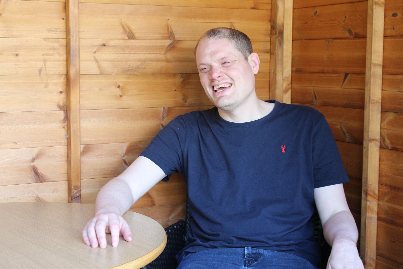 A white man wearing a black t-shirt sits in his summer house with a huge smile on his face.