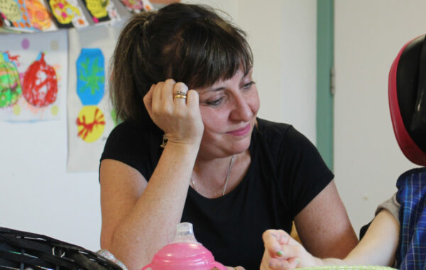 A white woman with dark hair and wearing a black t-shirt sits in an art room with her hand on her head. She is watching a client make art.