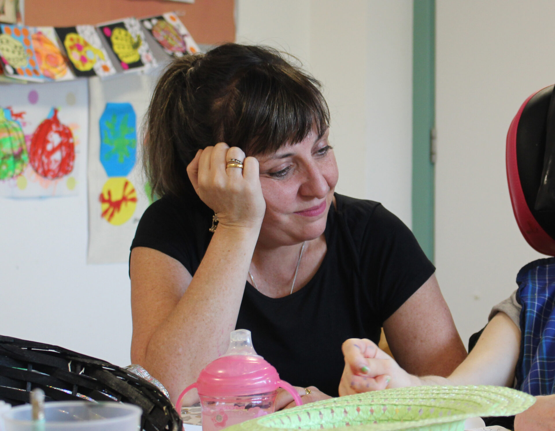 A white woman with dark hair and wearing a black t-shirt sits in an art room with her hand on her head. She is watching a client make art.
