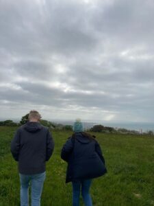 Two people - a man and a woman - are walking on the green downs. The sea is ahead of them and the clouds are moody.