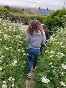 A woman with dark curly hair, wearing jeans and a grey hoodie, walks behind someone in a very grassy, shrubby path.