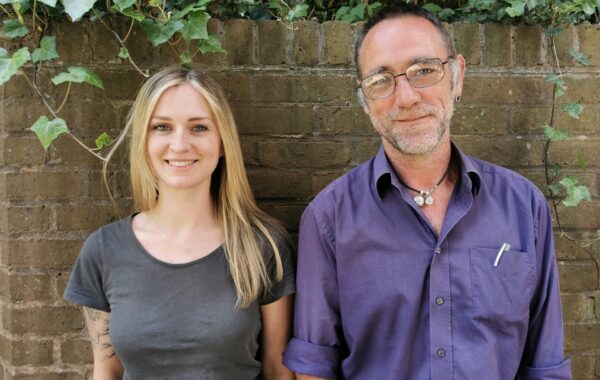 A woman with long blonde hair wears a grey t-shirt. She has a tattoo on her arm and is smiling broadly to the camera. Next to her is a man with a purple shirt and glasses. He is also smiling to the camera. They are both standing in front of a wall and ivy.