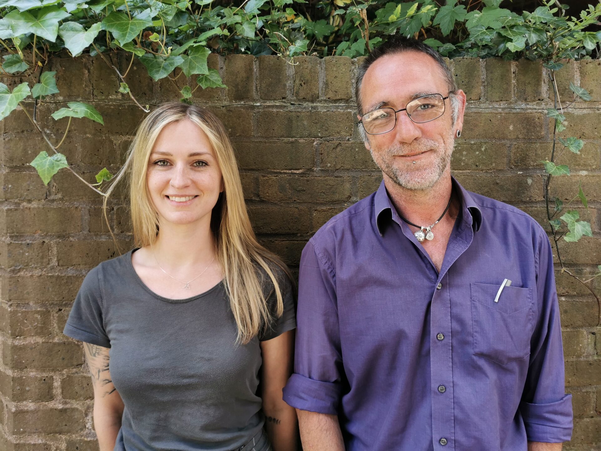 A woman with long blonde hair wears a grey t-shirt. She has a tattoo on her arm and is smiling broadly to the camera. Next to her is a man with a purple shirt and glasses. He is also smiling to the camera. They are both standing in front of a wall and ivy.