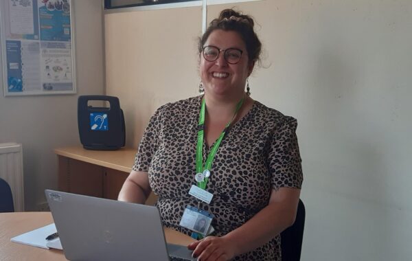 A very smiley woman with her hair in a bun is looking directly at the camera. She is in an office with her laptop open.