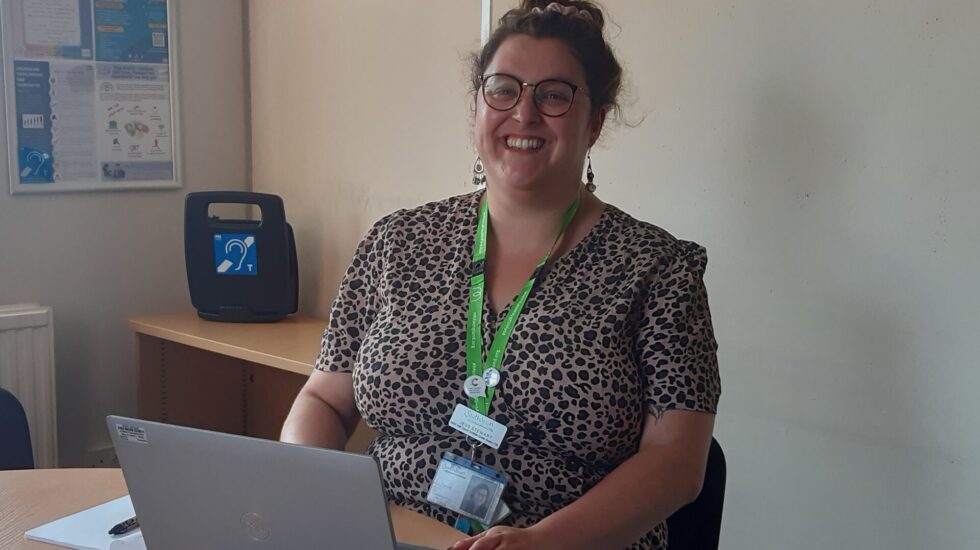 A very smiley woman with her hair in a bun is looking directly at the camera. She is in an office with her laptop open.