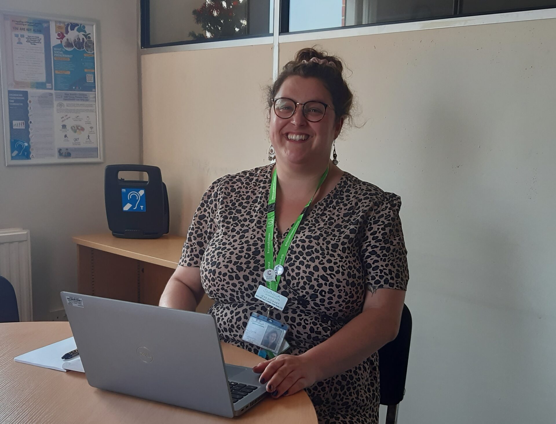 A very smiley woman with her hair in a bun is looking directly at the camera. She is in an office with her laptop open.