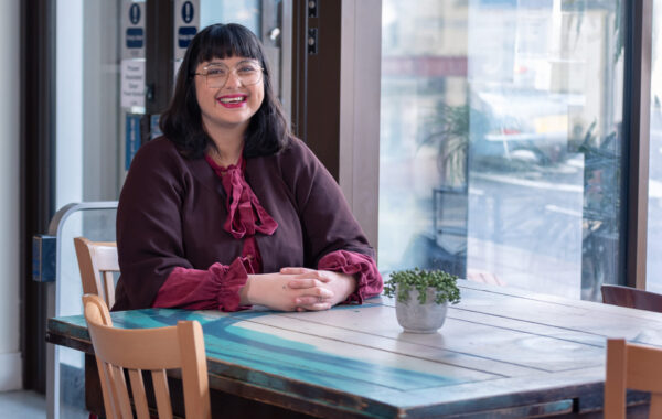 A woman with dark brown-black hair wearing glasses and a bright red lip, smiles broadly at the camera in a cafe