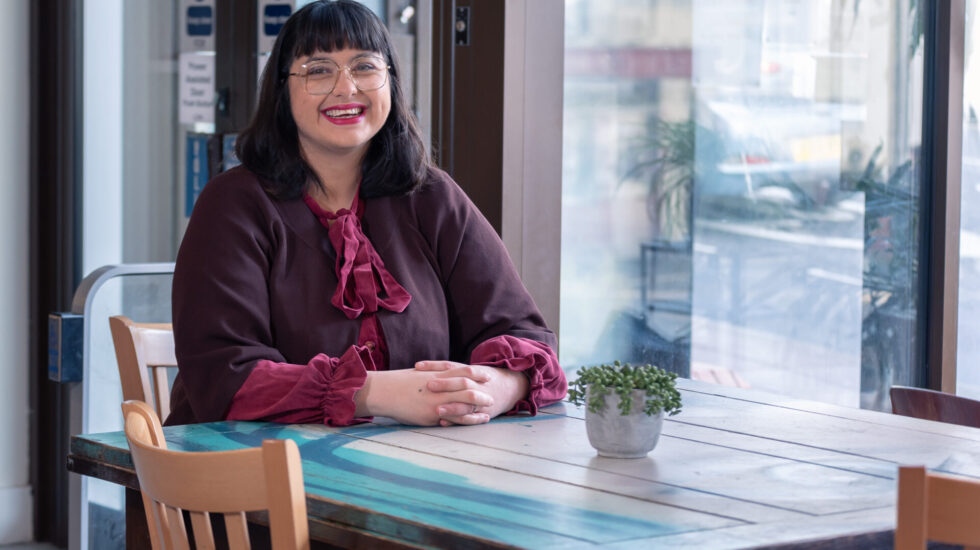 A woman with dark brown-black hair wearing glasses and a bright red lip, smiles broadly at the camera in a cafe