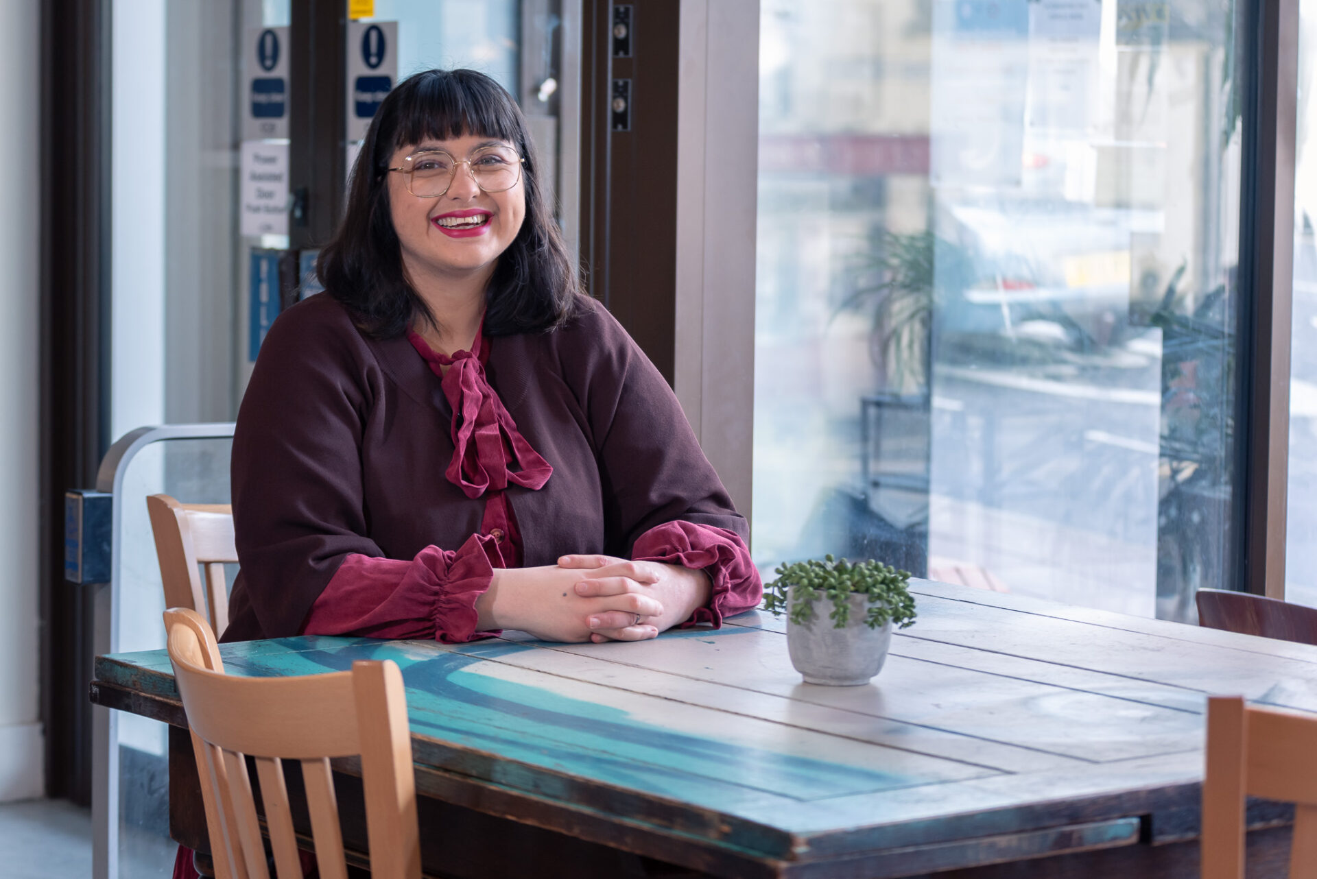 A woman with dark brown-black hair wearing glasses and a bright red lip, smiles broadly at the camera in a cafe