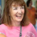 A white woman with auburn hair and wearing a pink t-shirt smiles broadly