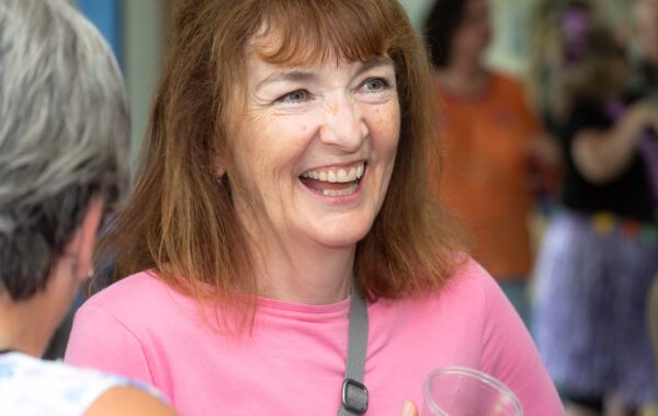 A white woman with auburn hair and wearing a pink t-shirt smiles broadly