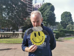 Fatboy Slim is in a park. He is looking directly to camera, holding a smiley face sheep mask