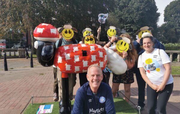 Fatboy Slim is in the foreground. Behind him is a red Shaun the Sheep sculpture and people with smiley face sheep masks