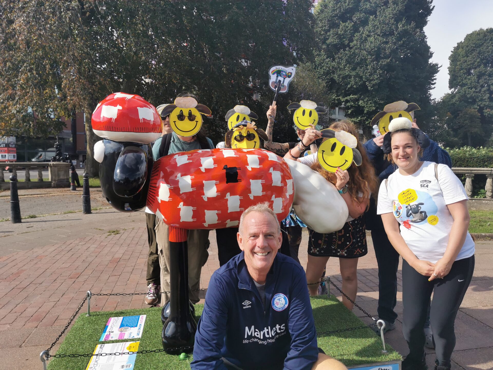 Fatboy Slim is in the foreground. Behind him is a red Shaun the Sheep sculpture and people with smiley face sheep masks