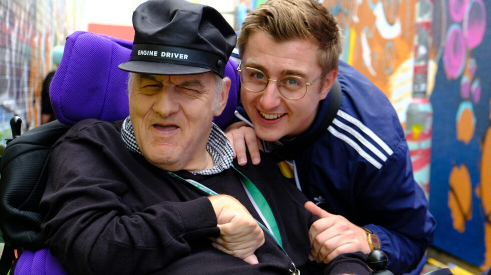 A Southdown Support Worker and a client in a wheelchair are smiling. They are in an alleyway with vibrant graffiti on the walls behind them.