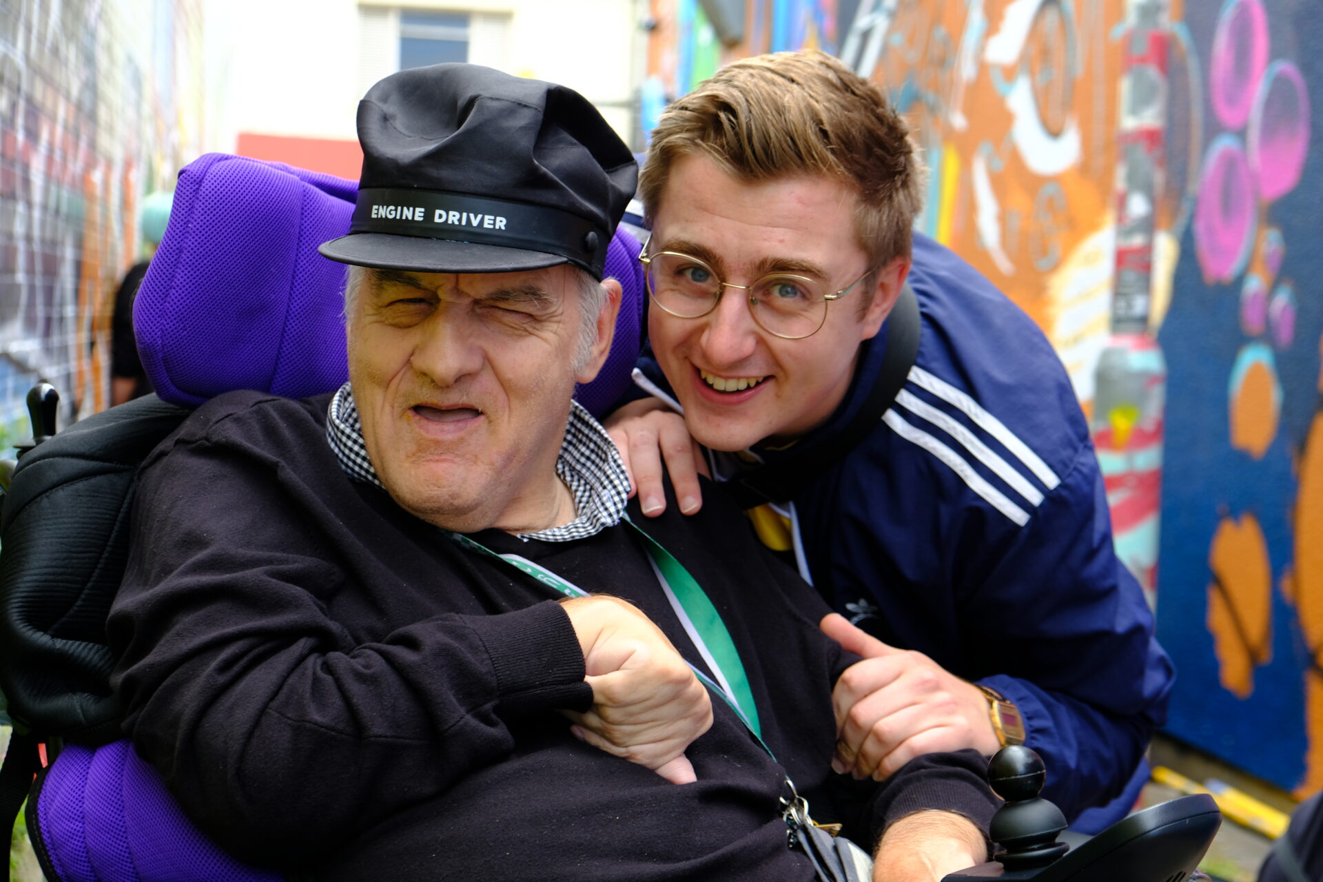 A Southdown Support Worker and a client in a wheelchair smiling. They are in an alleyway with vibrant graffiti on the walls behind them. 
