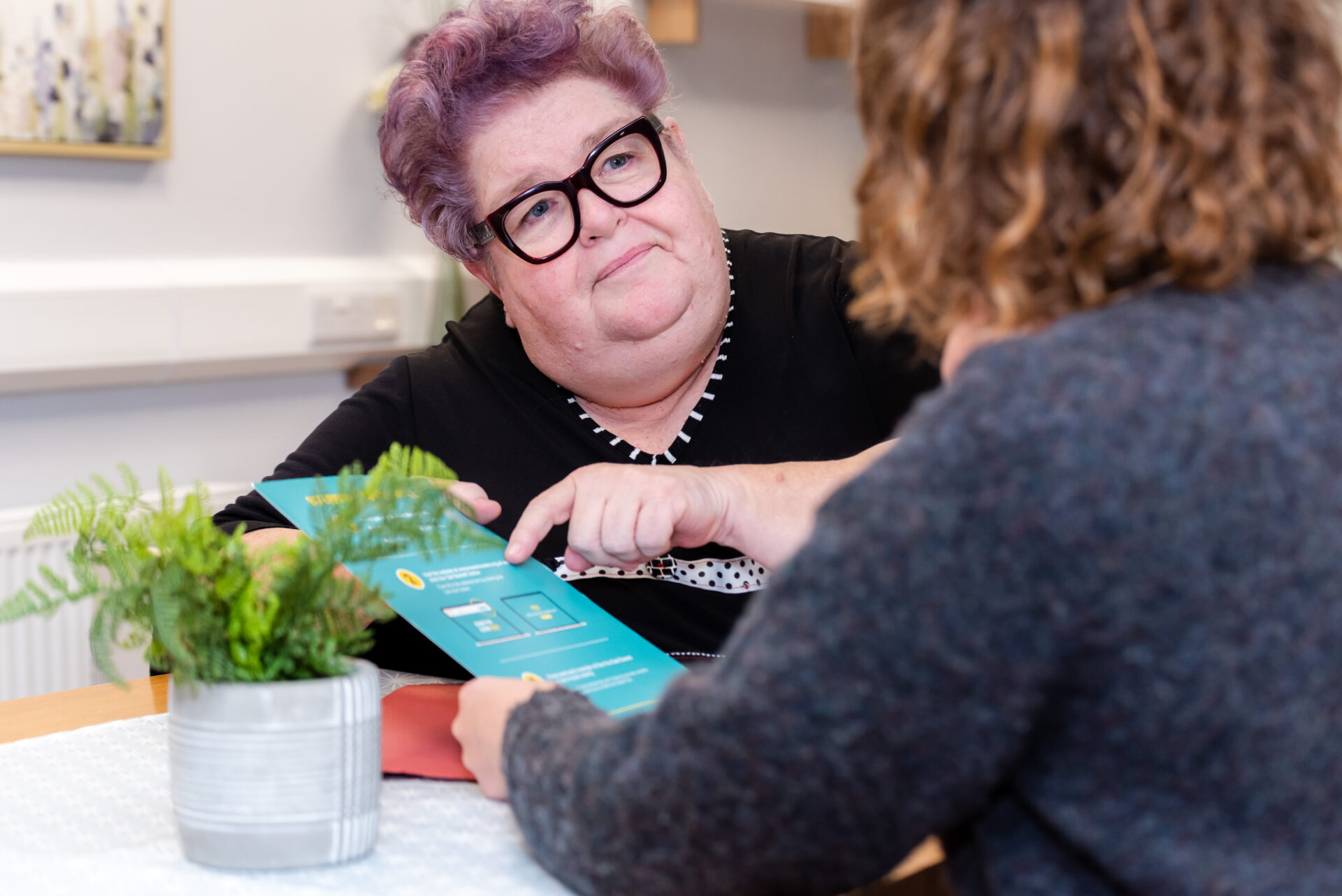 A white woman with black glasses and purple cropped hair is looking at a client with curly brown hair. She is holding a leaflet in her hands.