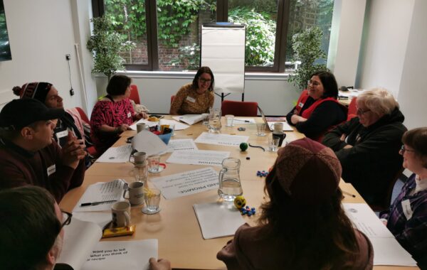 A group of people sit around a table with lots of paper and worksheets for a client voice workshop. They are all talking to each other.