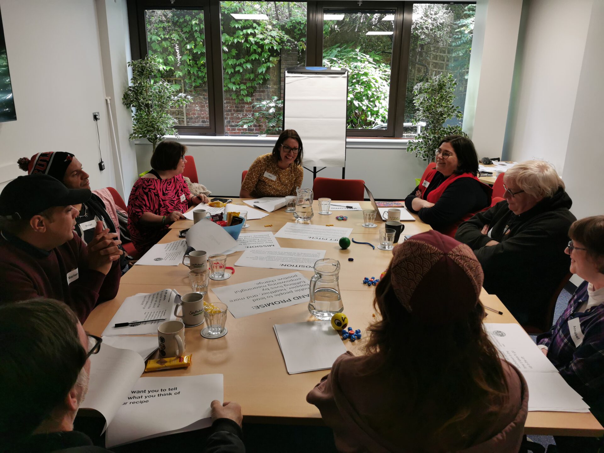 A group of people sit around a table with lots of paper and worksheets for a client voice workshop. They are all talking to each other.