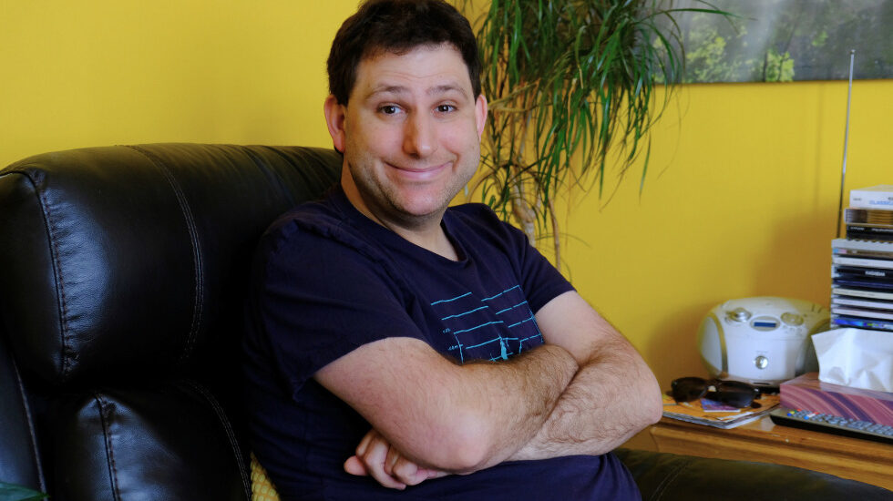 Southdown housing client smiling looking at the camera and crossing their arms. They are sitting on a leather sofa in a room with bright yellow walls.