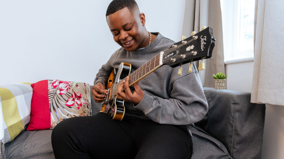 Southdown client is smiling whilst playing the guitar. They are sat on a sofa near the window.