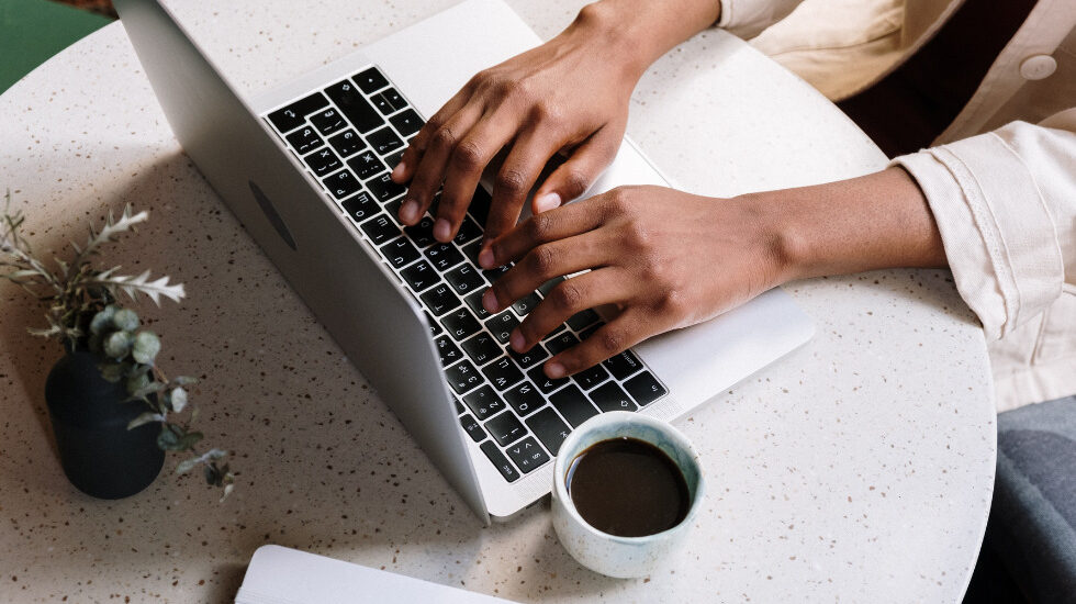 A person's hands are typing on their laptop on a table, with a black cup of coffee next to them.
