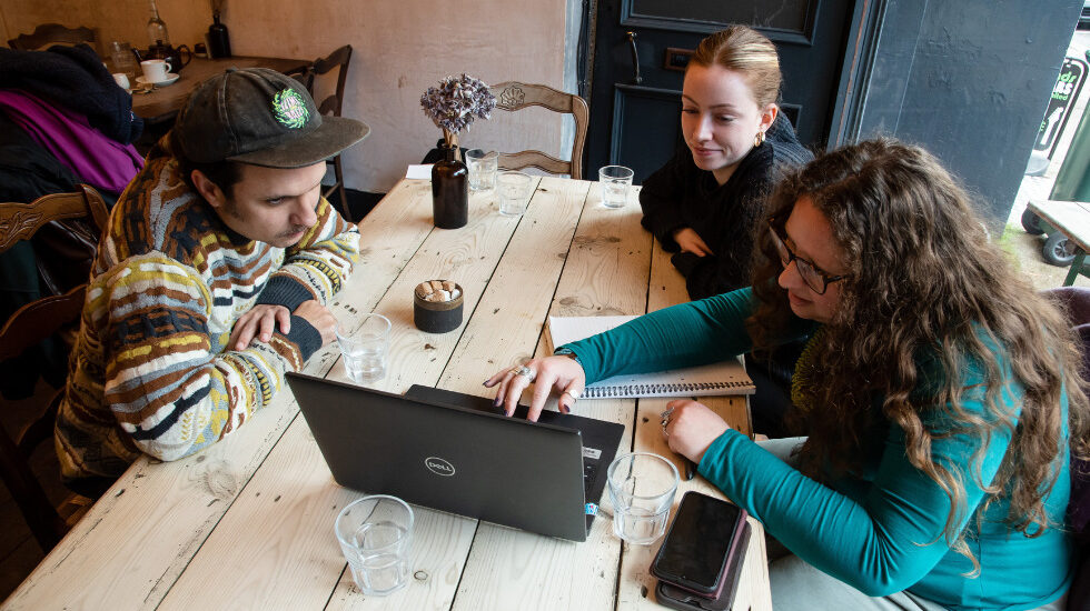 Three people are sat around a wooden table looking at a laptop.