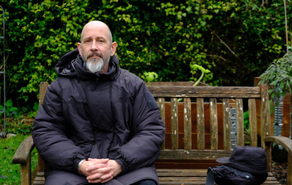 A white man with a smooth head and a grey beard is sitting on a garden bench. He is dressed in black and has a black bag next to him.