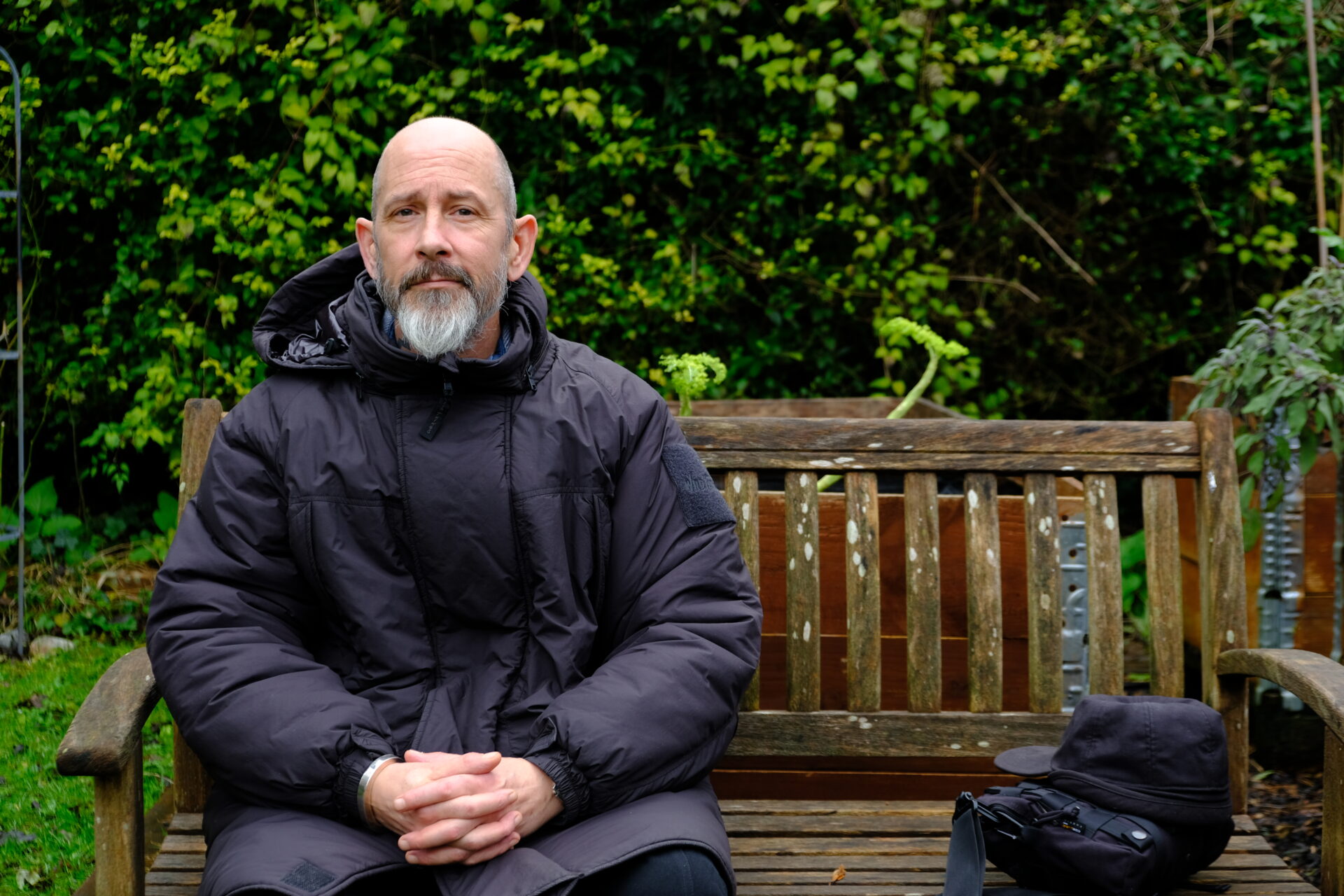 A white man with a smooth head and a grey beard is sitting on a garden bench. He is dressed in black and has a black bag next to him.