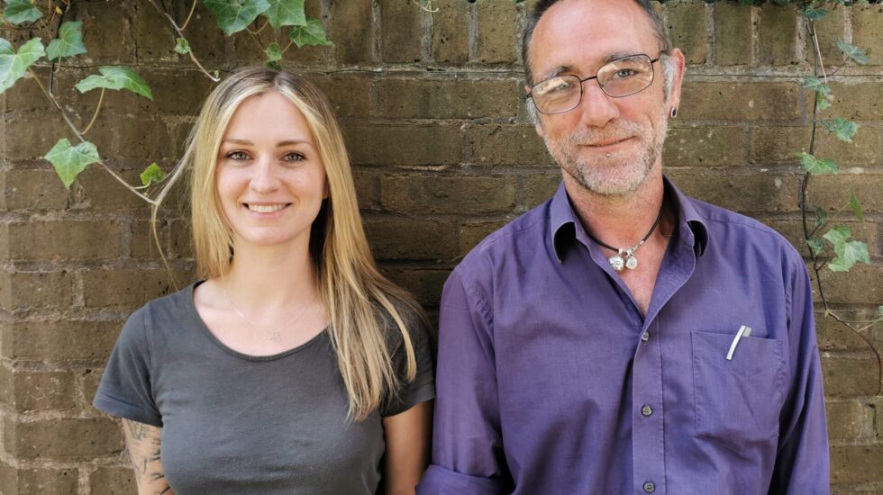 Two staff members standing next to each other smiling to the camera. The are outside and stood in front of a brick wall covered in ivy.