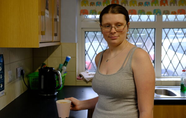 A young white woman with her hair tied back is standing in the kitchen. She is holding a white mug. The window is behind her.