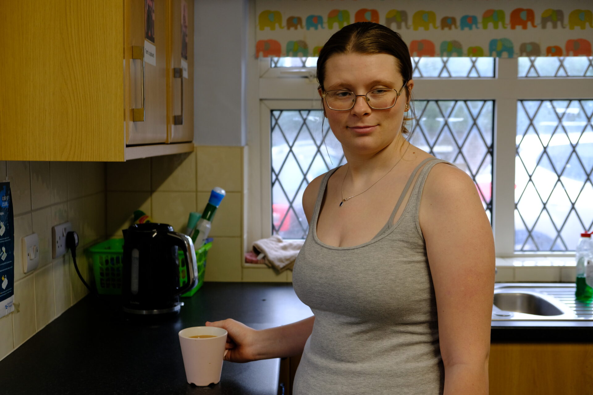 A young white woman with her hair tied back is standing in the kitchen. She is holding a white mug. The window is behind her.