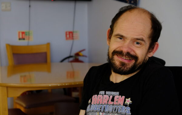 A white man with dark hair and a beard sits in his wheelchair in the kitchen. He is smiling at the camera.