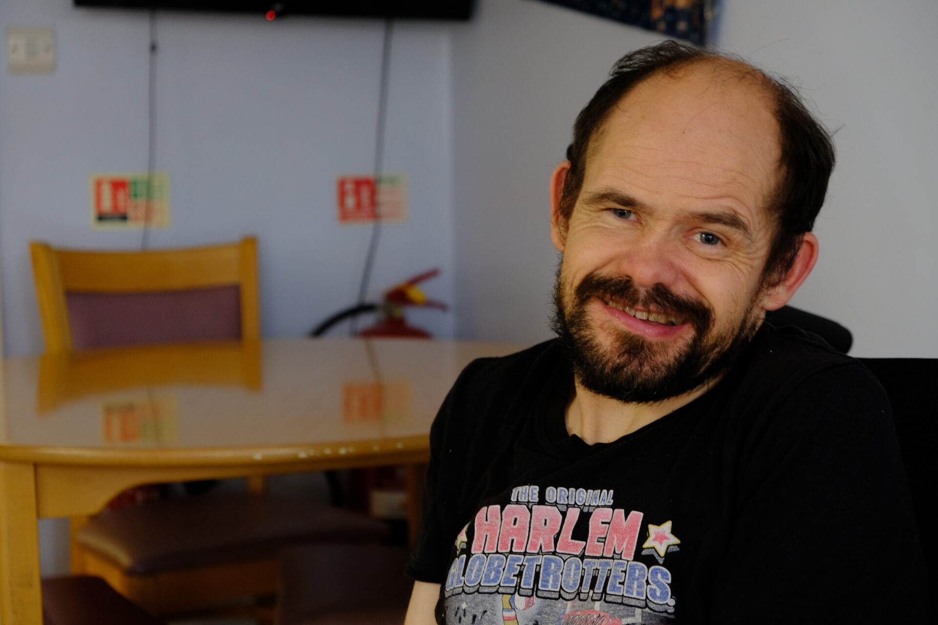 A white man with dark hair and a beard sits in his wheelchair in the kitchen. He is smiling at the camera.