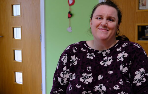 A white woman is smiling warmly to the camera. She is wearing a dark floral top and there are love hearts on the green wall behind her.