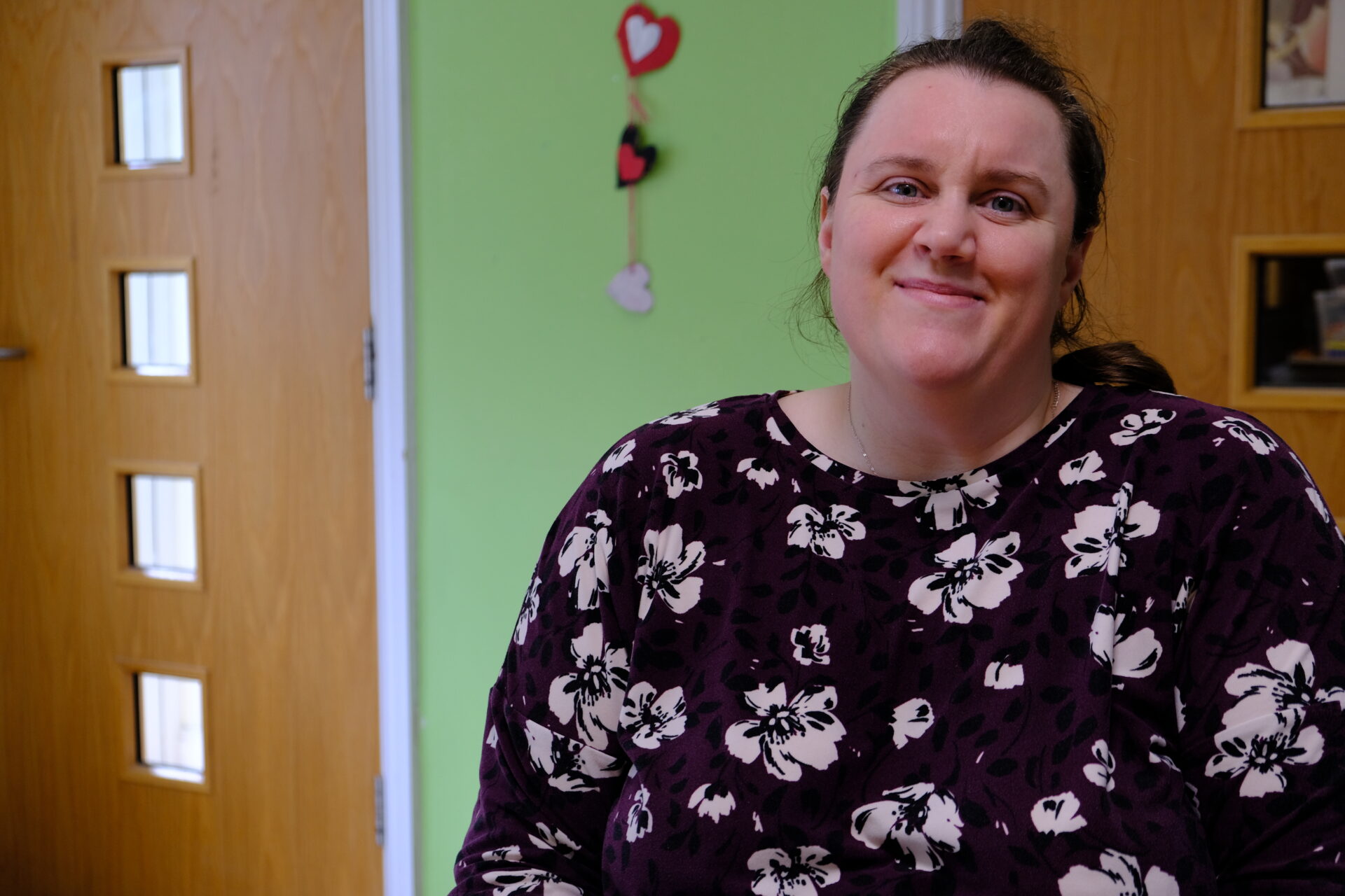 A white woman is smiling warmly to the camera. She is wearing a dark floral top and there are love hearts on the green wall behind her.