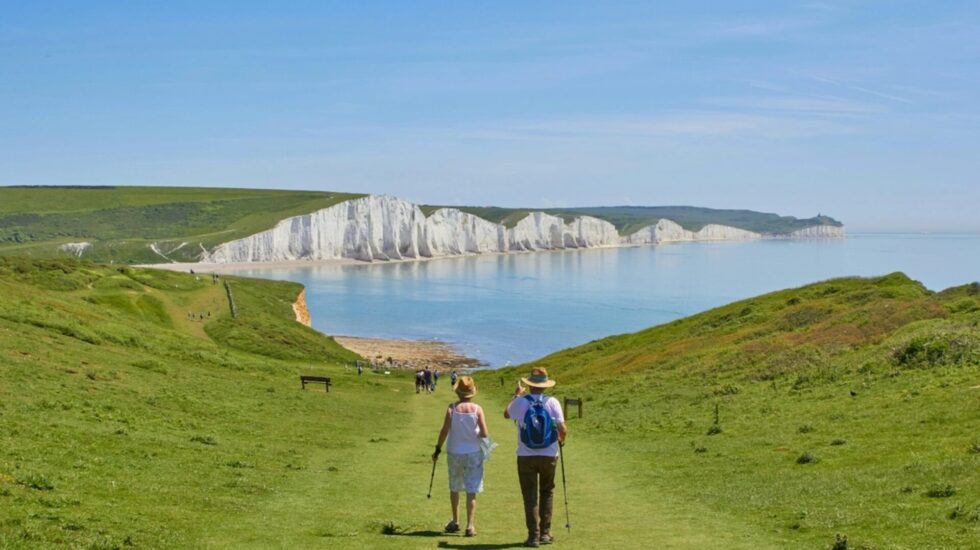 Two people are walking alongside each other on grass. In the background there are white cliffs in East Sussex.
