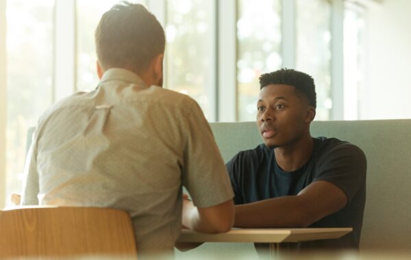 Two men are sat at a table talking to each other. There is sun shining through the window behind them.