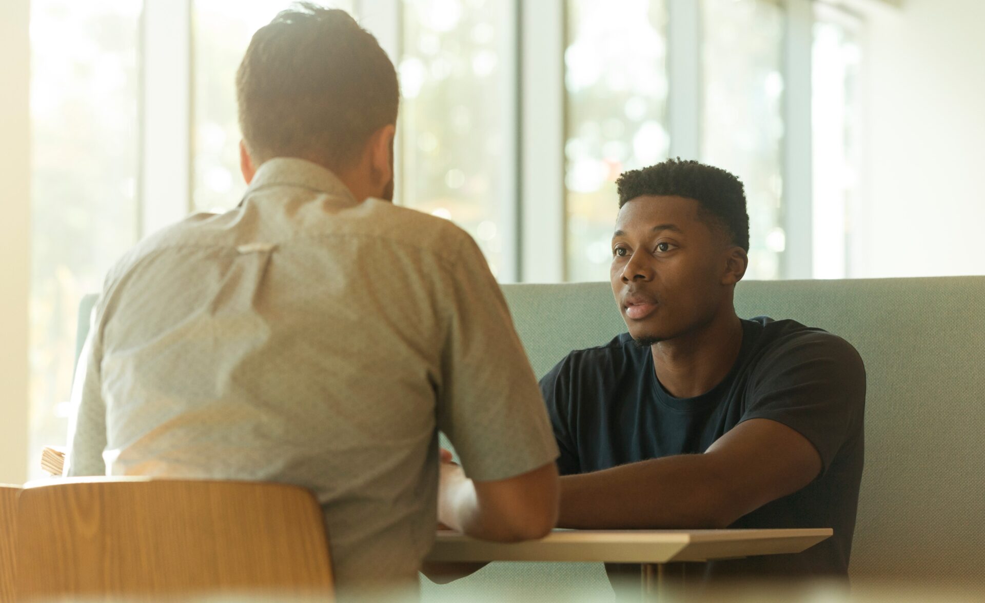 Two men are sat at a table talking to each other. There is sun shining through the window behind them.