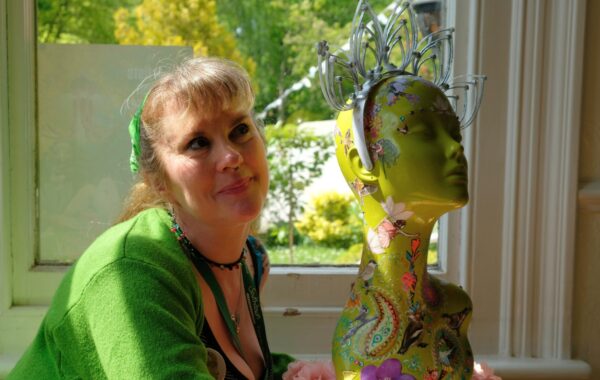 A woman is sat near a window, next to a mannequin head. It is decorated with artwork and is wearing a deocrative headband.