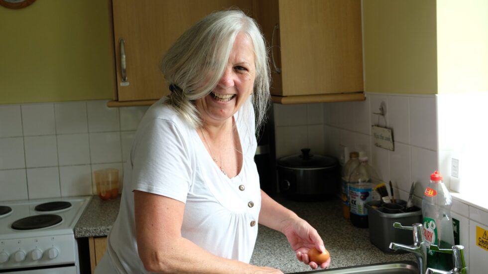 A woman with shoulder-length grey hair and light skin is standing in a kitchen, smiling warmly at the camera. She is wearing a white button-up t-shirt and holding an egg in her hand while washing it under the sink. The kitchen has wooden cabinets, a white tiled backsplash, and various kitchen items, including a slow cooker, bottles, and washing-up liquid, on the counter. The atmosphere is bright and homely.