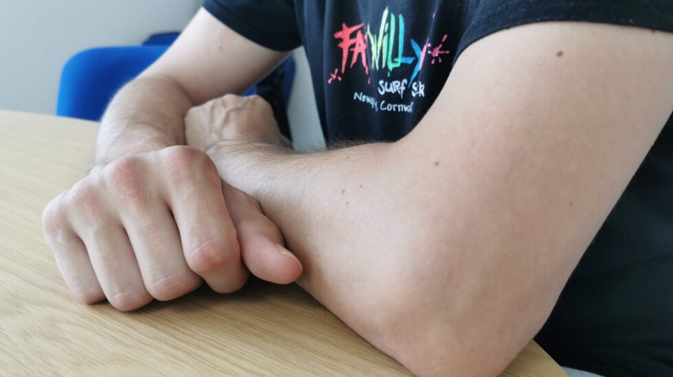 A close-up photograph of a person’s arms resting on a light wooden table. The individual is wearing a black T-shirt with a colourful "Family Surf" logo. The person is a client of the Pathways Home service, a Housing Support programme in West Sussex. The image conveys a sense of stability and support, reflecting the service's goal of helping individuals build independence through responsive housing support.