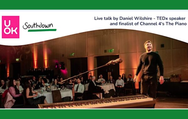 Promotional banner for a live talk on mental health awareness by Daniel Wilsher, TEDx speaker and finalist of Channel 4’s The Piano. The image shows Daniel on stage in front of a seated audience, with a microphone and keyboard in the foreground. The UOK and Southdown logos are displayed at the top of the banner. The talk's theme is - Building community through service.