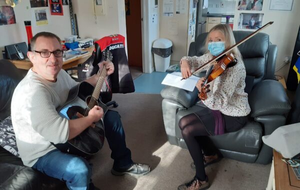 A Southdown client with a learning disability sits on a sofa playing an acoustic guitar and smiling at the camera. Opposite him, Anita Beverton, Southdown’s Head of Learning Disability, wears a face mask and floral blouse while playing the violin. They are seated in a cosy, personalised living space filled with sports memorabilia, posters, and certificates, sharing a music practice session that reflects connection, confidence, and creativity.
