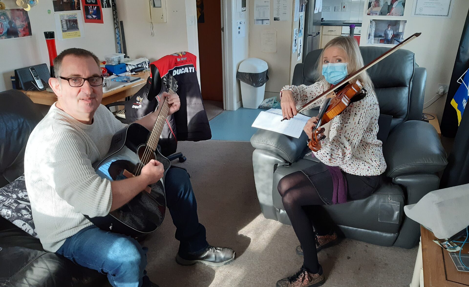 A Southdown client with a learning disability sits on a sofa playing an acoustic guitar and smiling at the camera. Opposite him, Anita Beverton, Southdown’s Head of Learning Disability, wears a face mask and floral blouse while playing the violin. They are seated in a cosy, personalised living space filled with sports memorabilia, posters, and certificates, sharing a music practice session that reflects connection, confidence, and creativity.