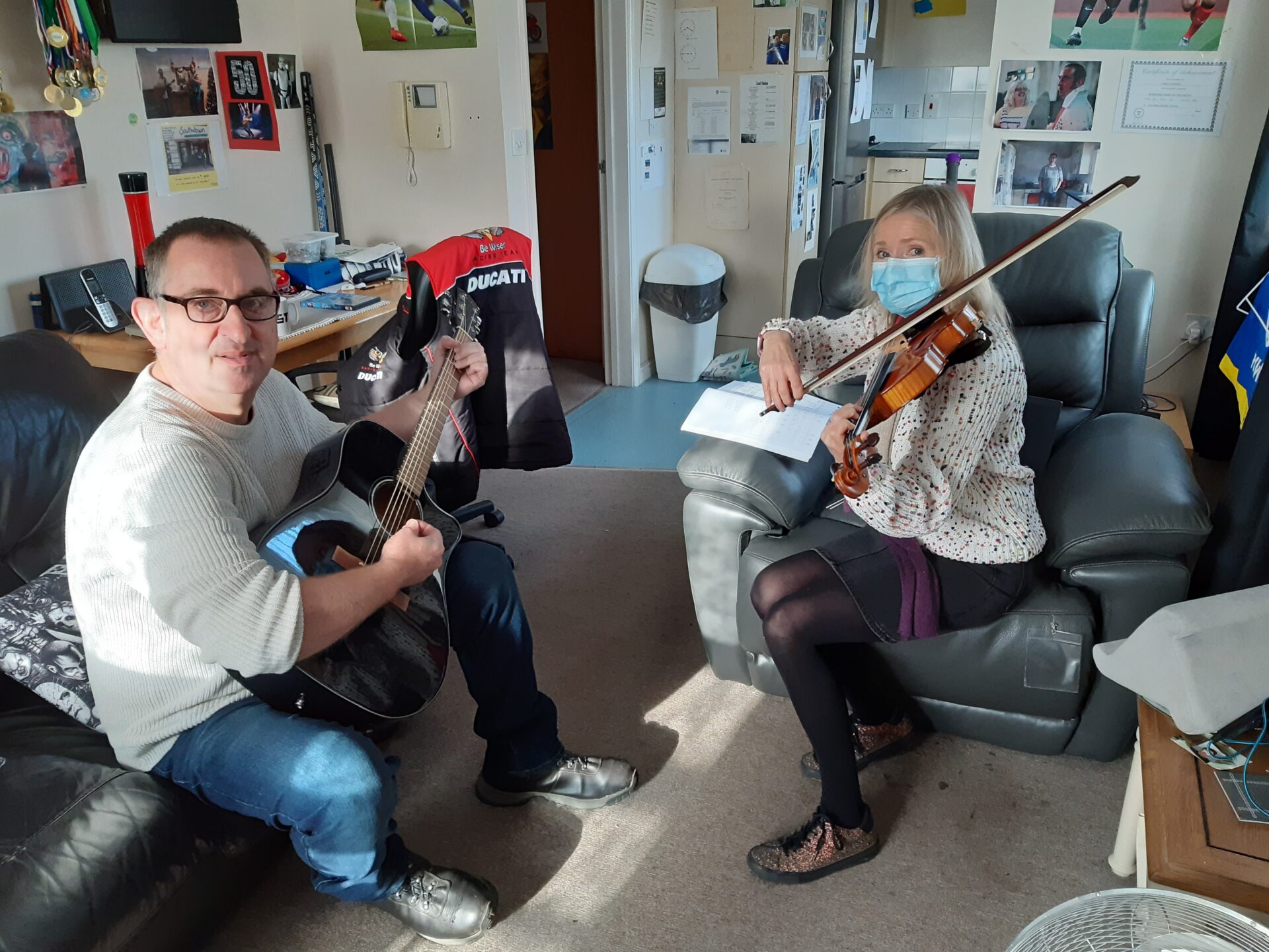 A Southdown client with a learning disability sits on a sofa playing an acoustic guitar and smiling at the camera. Opposite him, Anita Beverton, Southdown’s Head of Learning Disability, wears a face mask and floral blouse while playing the violin. They are seated in a cosy, personalised living space filled with sports memorabilia, posters, and certificates, sharing a music practice session that reflects connection, confidence, and creativity.