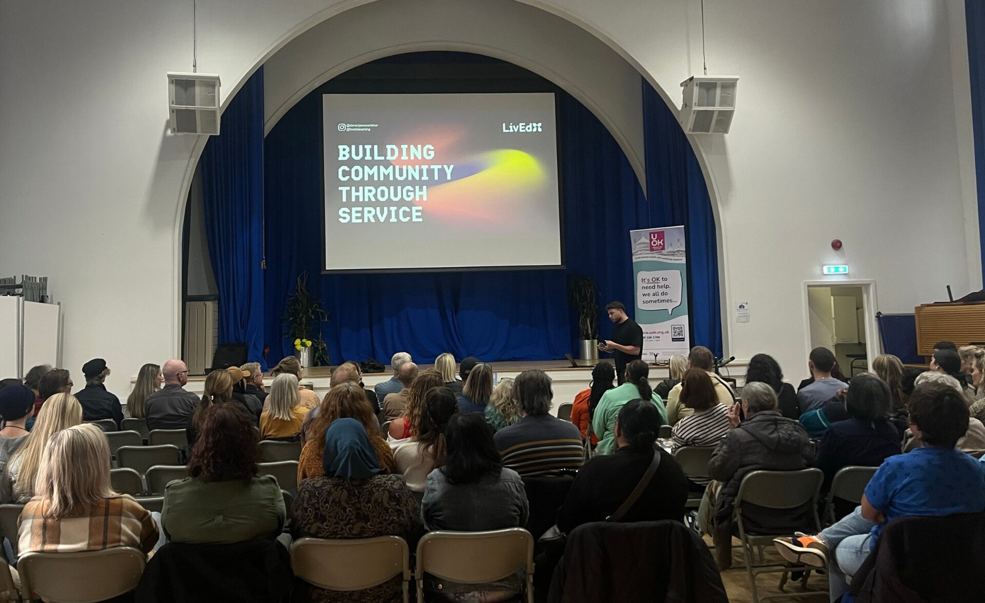 A mental health talk in progress, delivered by Daniel Wilsher, to a seated audience in a community hall. A large screen behind him reads 'Building Community Through Service' and a UOK banner stands nearby, displaying the message 'It's OK to need help, we all do sometimes.'