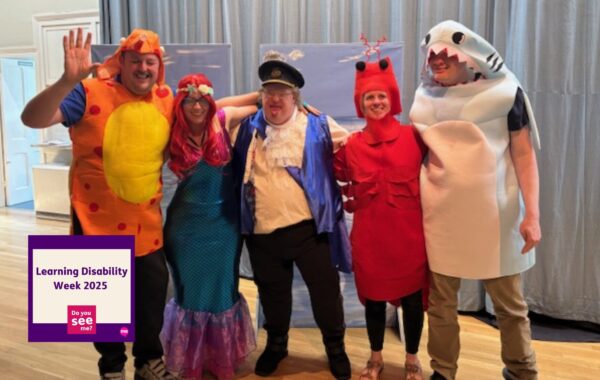 A group of five people from the Milchester Players theatre group, including clients and Support Workers from an East Sussex service, are dressed in colourful sea-themed costumes on stage. They are smiling and posing together in outfits such as a fish, mermaid, ship captain, lobster, and shark. A "Learning Disability Week 2025 – Do You See Me?" badge appears in the corner of the image.
