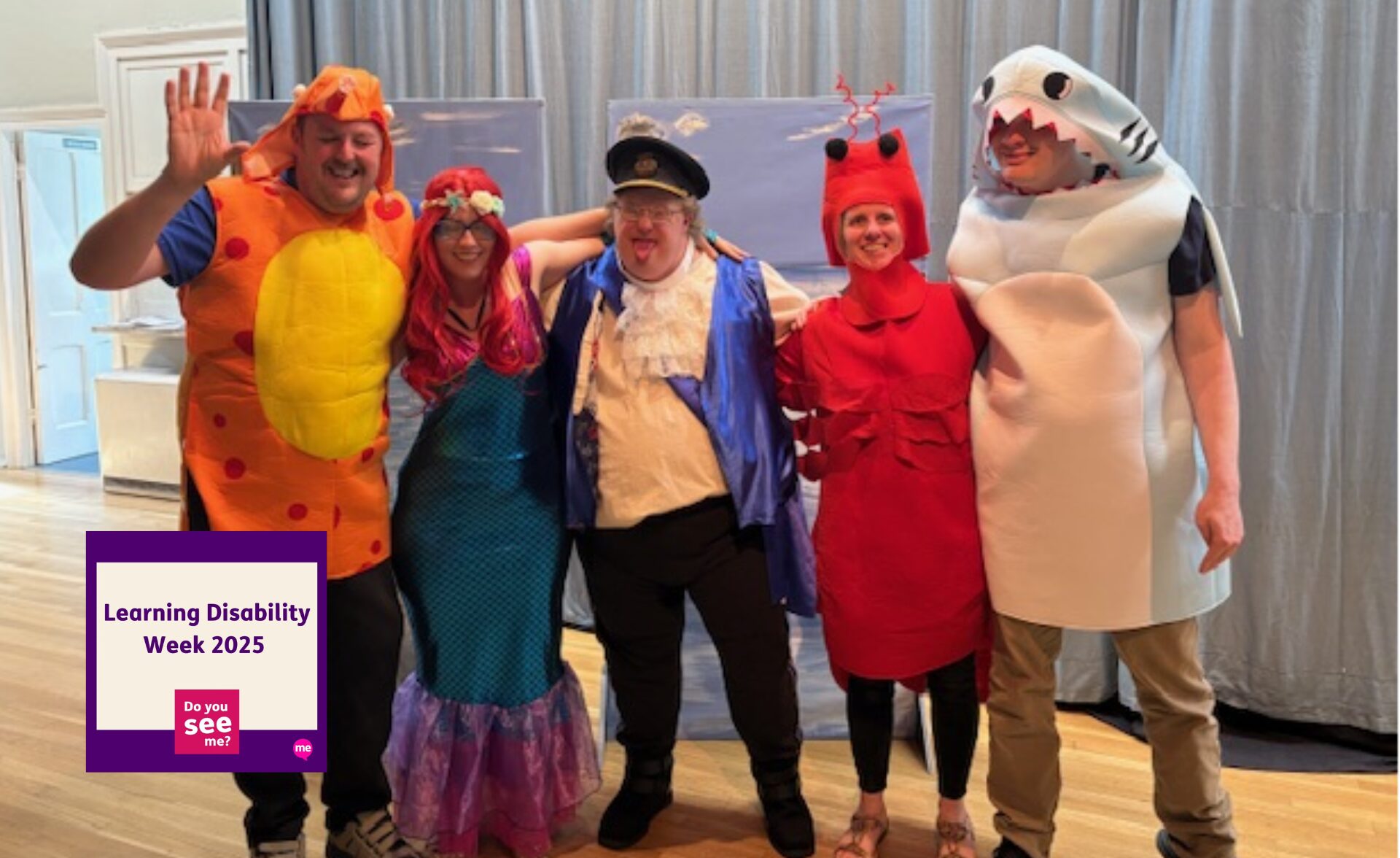 A group of five people from the Milchester Players theatre group, including clients and Support Workers from an East Sussex service, are dressed in colourful sea-themed costumes on stage. They are smiling and posing together in outfits such as a fish, mermaid, ship captain, lobster, and shark. A "Learning Disability Week 2025 – Do You See Me?" badge appears in the corner of the image.