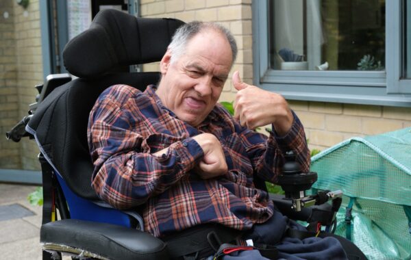 A middle-aged man with grey hair, wearing a red and navy checked shirt, sits in a powered wheelchair outside a supported living residence. He is smiling warmly and giving a thumbs-up with his right hand, expressing confidence and positivity. The setting includes a brick wall, large windows, and a green mini greenhouse, suggesting a homely and supportive environment. The man is a client of Southdown’s learning disability service